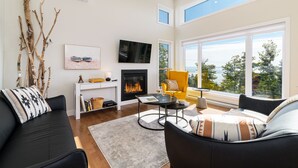 Una sala de estar moderna con chimenea, un sofá, un sillón amarillo y un ventanal con vistas a árboles y al cielo.
