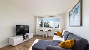 Una sala de estar moderna con un televisor de pantalla plana, un sofá gris con cojines amarillos, un escritorio blanco y un ventanal con vistas a árboles y al cielo.