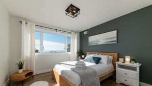 Un dormitorio con cama de madera, ropa de cama blanca, vista al mar a través de ventanas grandes, un cuadro en la pared y una mesita de noche con una lámpara.