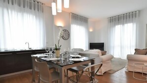 A modern living room with a dining area, featuring a glass table, metal chairs, a sofa, and a TV.