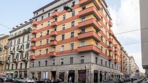 A multi-story building with balconies, a ground-level entrance, and a street view with parked cars.
