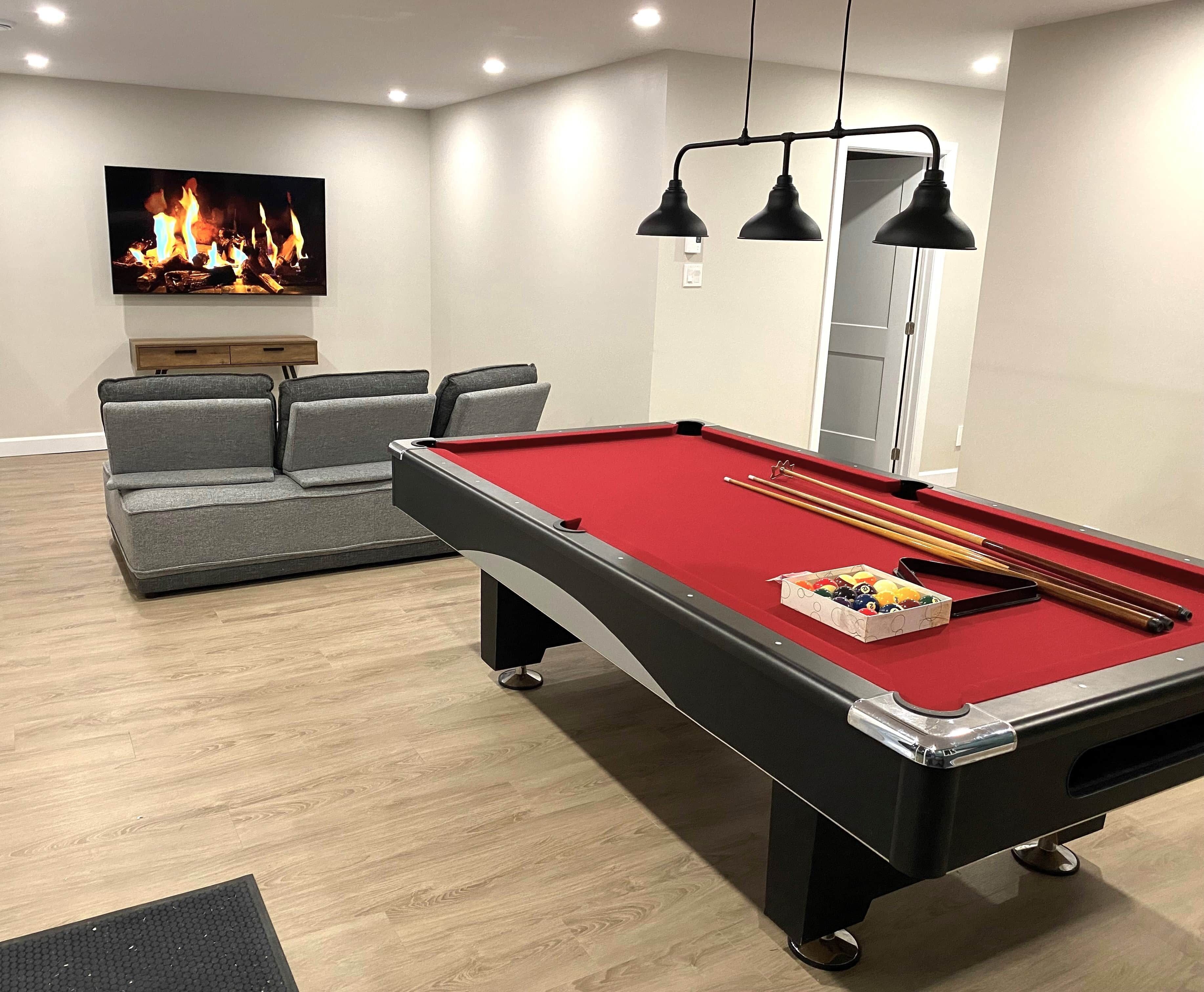 A pool table with a red felt surface, a set of pool cues, and a box of billiard balls. There is a grey sofa with cushions in the background and a painting of a fire on the wall.