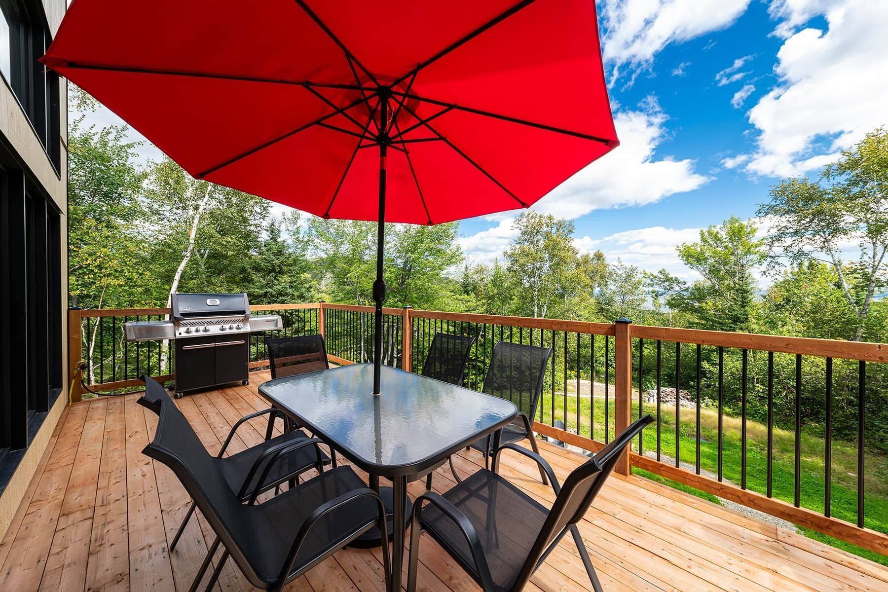 A deck with a glass table and chairs, a red umbrella, and a barbecue grill.