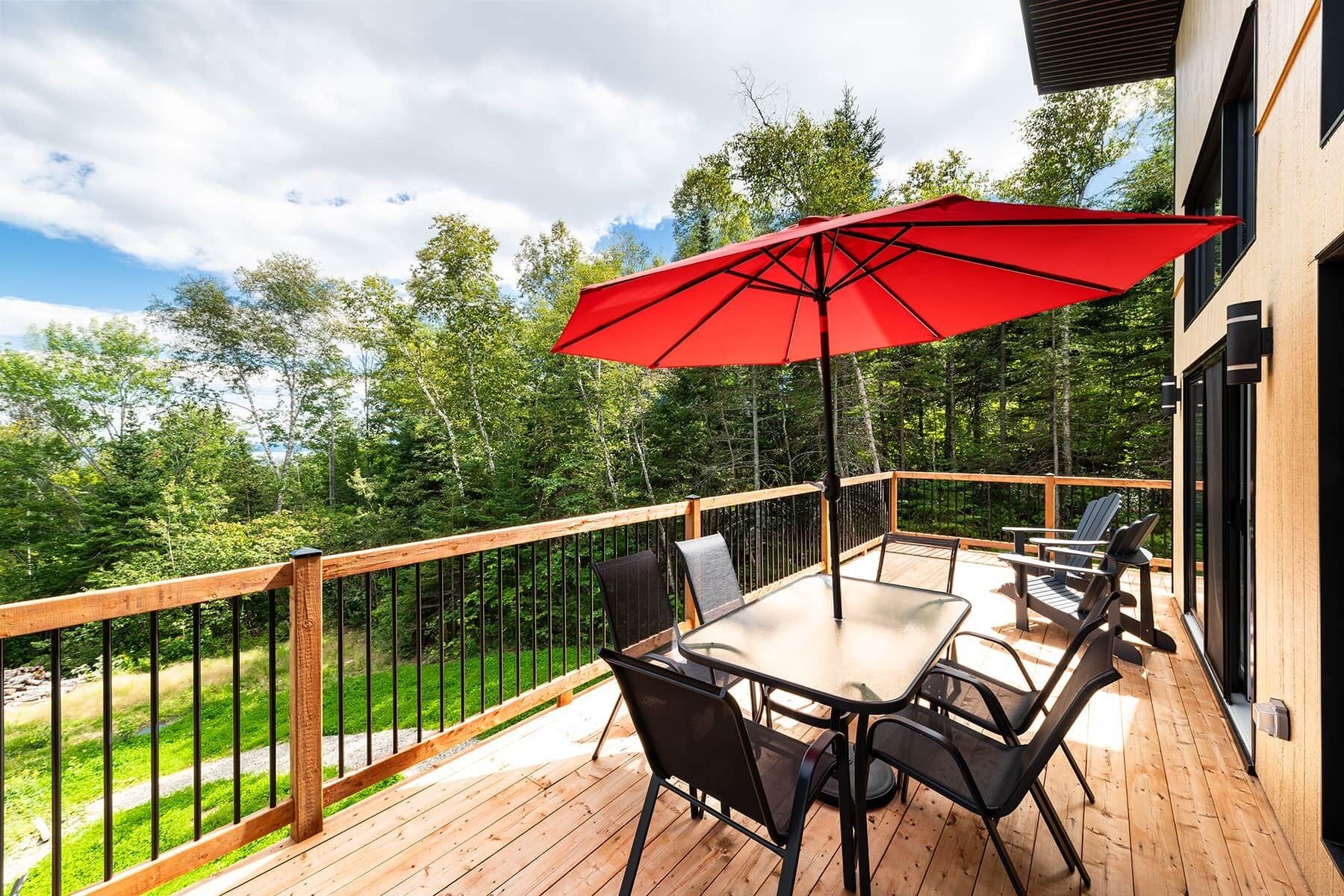 Une terrasse en bois avec une table et des chaises, un parasol rouge et une vue sur les arbres.