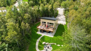 A modern house with a deck, surrounded by lush greenery and a gravel path. - Hôtel à la maison - Chez Fugère, spa et billard (Les Éboulements)