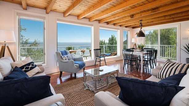 Ein geräumiges Wohnzimmer mit Holzboden, großen Fenstern mit Blick ins Freie, einem Essbereich mit Tisch und Stühlen sowie einer gemütlichen Sitzecke mit Sofa und Sesseln.
