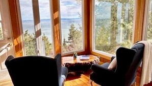 A cozy cabin interior with wooden floors, large windows offering a view of a lake and forest, and two black armchairs. - Hôtel à la maison - Villa du Cap Blanc, spa et Vue (Saint-Irénée)