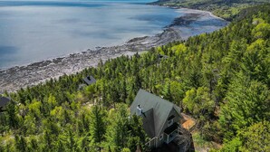 A house with a sloped roof, surrounded by dense green forest, overlooking a rocky shoreline and calm sea. - Hôtel à la maison - Villa du Cap Blanc, spa et Vue (Saint-Irénée)