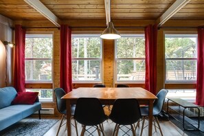 Et hyggeligt rum med træloft, et spisebord med stole, en blå sofa med rød pude og store vinduer med røde gardiner.