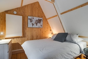 Et soveværelse med skråvæg, trævægge, en seng med gråt og hvidt sengetøj, et skrivebord med lampe og et verdenskort på væggen.