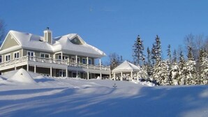 Une grande maison à deux étages, dotée d’un balcon et d’un belvédère, entourée d’arbres couverts de neige.