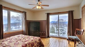 Ein Schlafzimmer mit Bett, Deckenventilator, Schminktisch und Blick auf eine verschneite Landschaft durch große Fenster.