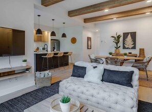 Sala de estar moderna com sofá cinzento, televisão de tela plana, área de cozinha com banquetas e mesa de jantar com cadeiras de vime.