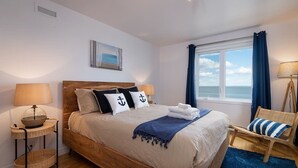 Un dormitorio con cama de madera, dos mesitas de noche con lámparas, vista al mar a través de una ventana y una silla con cojín a rayas.