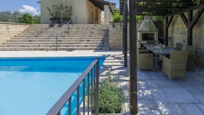 Una piscina con deck de piedra, un espacio comedor al aire libre techado con mobiliario de mimbre, y una escalera de piedra que lleva a un edificio.