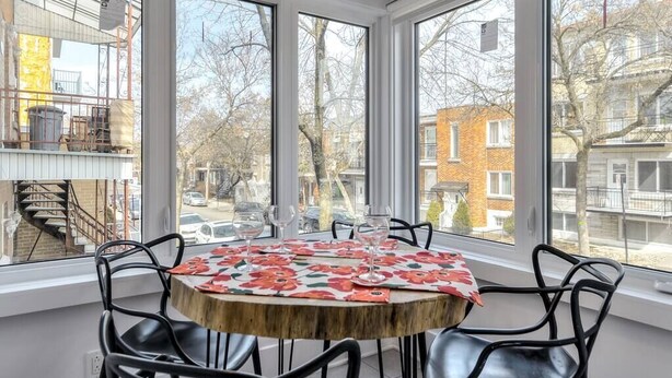 Una habitación con una mesa redonda y cuatro sillas, ventanas amplias con vistas a edificios y árboles, y una escalera visible a través del ventanal.