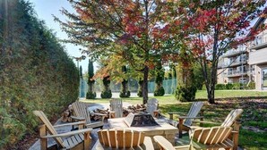 An outdoor seating area with Adirondack chairs and a fire pit, surrounded by trees and a hedge.