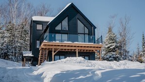 Moderni, kaksikerroksinen mökki, jossa on puinen terassi ja suuret ikkunat. Mökin ympärillä on lumipeitteisiä puita.