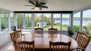 A dining area with a large oval table and six wooden chairs, large windows offering a view of a lake and greenery, and a ceiling fan.