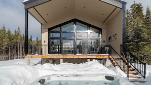 A modern cabin with a large glass window, a wooden deck, and a hot tub in the snow. - Hôtel à la maison - Le Lys, nature et spa (Shannon)