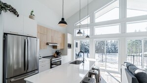A modern kitchen with a large island, stainless steel appliances, and a view of a snowy deck through large windows. - Hôtel à la maison - Le Lys, nature et spa (Shannon)