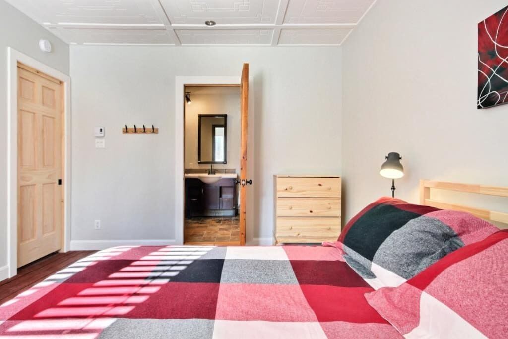 A bedroom with a checkered bedspread, a wooden dresser, a nightstand with a lamp, a door leading to a bathroom, and a kitchen visible through an open door.
