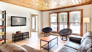 Ein modernes Wohnzimmer mit Holzboden, einem Flachbildfernseher, Ledersofas und einem großen Fenster mit Blick auf Bäume.