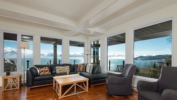 A spacious living room with a large black leather sofa, a wooden coffee table, and a grey armchair. The room has large windows offering a view of the sea and mountains.