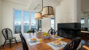 A dining area with a wooden table, black chairs, a bowl of fruit, croissants, and orange juice. There is a fireplace and a view of the outdoors.