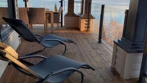 A covered outdoor area with wooden flooring, two lounge chairs, a small table, and a fireplace.