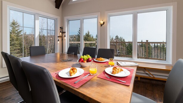 Una mesa de desayuno dispuesta con cruasanes, fruta y jugo de naranja en una habitación con ventanas grandes y vista a árboles.
