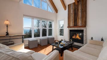 A spacious living room with a fireplace, large windows, and a high ceiling with exposed beams.