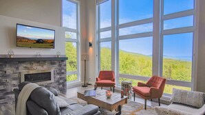A spacious living room with a fireplace, large windows, and a television mounted on the wall. - Hôtel à la maison - Panorama, spa et fleuve (Les Éboulements)
