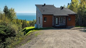 A single-story house with a stone facade and a wooden extension, situated on a gravel driveway with a view of the ocean. - Hôtel à la maison - Panorama, spa et fleuve (Les Éboulements)