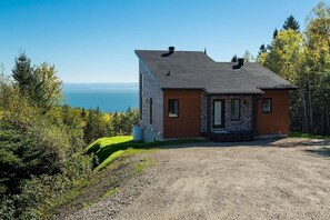 Une maison de plain-pied avec une façade en pierre et une extension en bois, située sur une allée de gravier et offrant une vue sur l’océan.