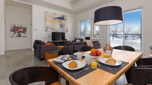 Una sala de estar moderna con comedor, que cuenta con un gran ventanal, una lámpara colgante negra y un cuadro en la pared.