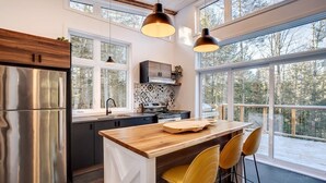 A modern kitchen with a wooden island, stainless steel appliances, and large windows offering a view of the outdoors. - Hôtel à la maison - Chalet Kölega, spa et nature (Potton)