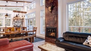 A spacious living room with a fireplace, a dining area, and a large window overlooking a forest. - Hôtel à la maison - Chalet Kölega, spa et nature (Potton)