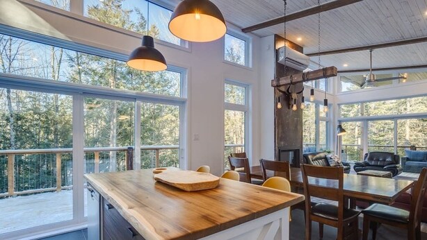 A spacious kitchen with a wooden island, large windows, and a dining area with chairs and a sofa. - Hôtel à la maison - Chalet Kölega, spa et nature (Potton)