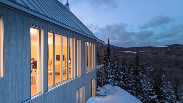 A modern building with large windows, a metal roof, and a view of a snowy landscape.