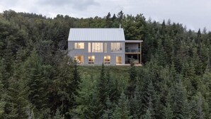 A modern two-story house with large windows and a balcony, surrounded by dense forest.