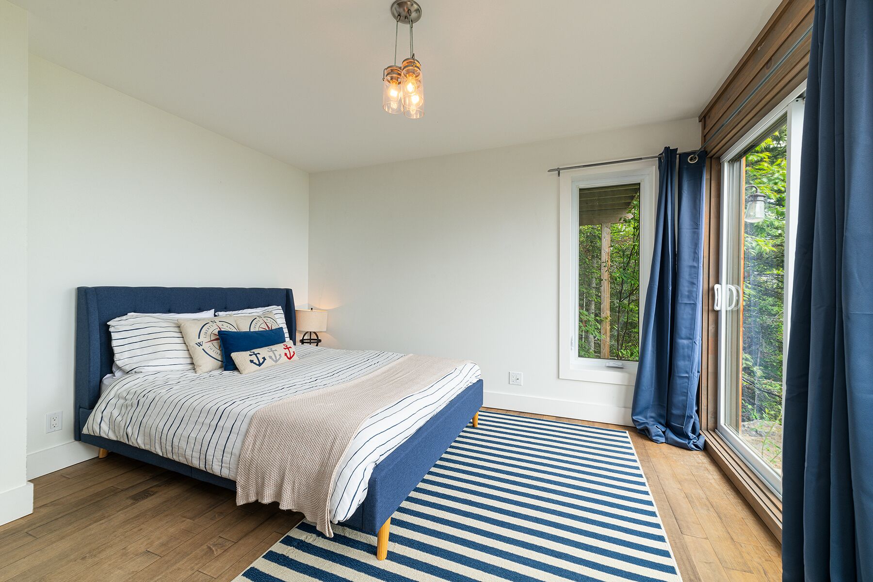 Een slaapkamer met een bed, een nachtkastje, een lamp, een raam met gordijnen en uitzicht op groen buiten.