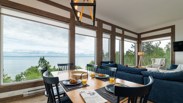 A dining area with a wooden table, chairs, and a view of a lake. - Hôtel à la maison - Le Phare d'Irène spa et fleuve (Saint-Irénée)