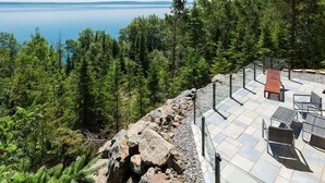 A patio with outdoor furniture overlooking a forest and a body of water. - Hôtel à la maison - Le Phare d'Irène spa et fleuve (Saint-Irénée)