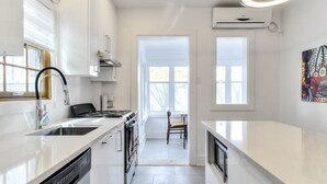 A modern kitchen with white cabinetry, a dishwasher, a stove, and a sink. There is a window providing natural light and a door leading to another room. - Hôtel à la maison - Le Gaillard (Montreal)