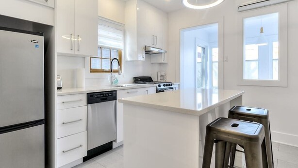 A modern kitchen with white cabinetry, a stainless steel refrigerator, a dishwasher, a gas stove, and a large island with stools. - Hôtel à la maison - Le Gaillard (Montreal)