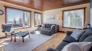 Ein geräumiges Wohnzimmer mit Holzbalkendecke, großen Fenstern mit Blick ins Freie, einem grauen Sofa, einem grauen Sessel und einem kleinen Couchtisch.