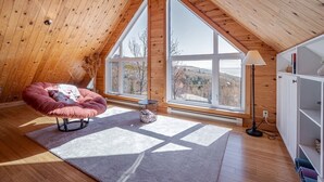Ein geräumiges Dachzimmer mit Holzbalken, großen Fenstern, einem rosafarbenen Sessel und einer weißen Lampe.