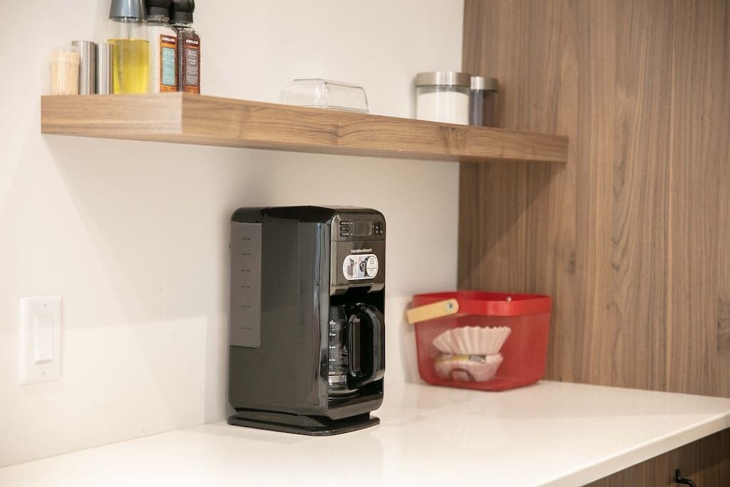 Un coin cuisine avec une machine à café posée sur un plan de travail blanc, une étagère en bois au-dessus contenant divers ustensiles de cuisine, et un récipient rouge avec une anse jaune.