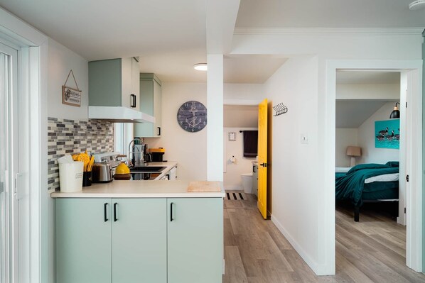 Una cocina compacta con salpicadero de azulejos, un reloj y una puerta amarilla que da a un dormitorio con cama y un cuadro.